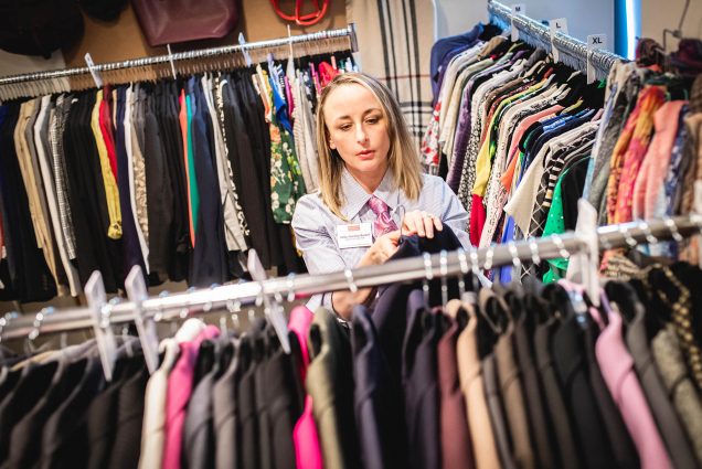 Photo: A white woman with shoulder length blonde hair is featured in the center of this photo, picking out clothes from a clothing rack in front of her. There are other clothing racks, one in the right hand side of the photo and behind. She is wearing a blue button up with a red tie and a name tag on her left-hand side.