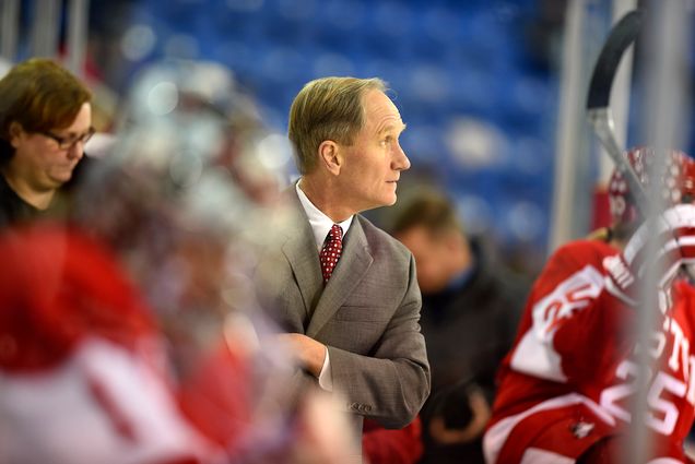 Photo: Brian Durocher, a white man wearing a tan suit, looks off to the right during a women's ice hockey game. He stands with arms crossed as he leans to the right to get a closer look.