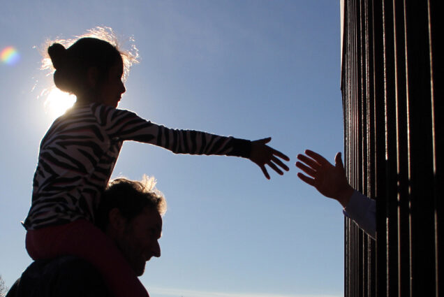 Photo: A girl from Anapra, a neighbourhood on the outskirts of Ciudad Juarez in Mexico, touches hands with a person on the United States through the border fence, during a prayer with priests and bishops from both countries to ask for the migrants and people of the area, on February 26, 2019. Silhouetted by the sun, a young girl sits on the shoulders of an adult as she reaches towards a shadow hand reaching through a fence.