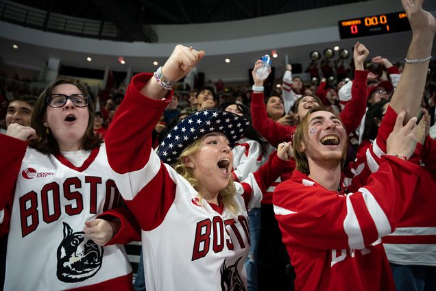 Photo: BU Students wearing large white and red "Boston" ice hockey jerseys cheer and celebrate BU men's hockey in the crowd during a game. Students from left to right: Taelor Anderson(CAS'24), Riley Egan (COM'25), Evan Teplensky(CAS'23).