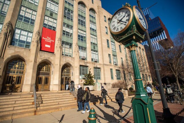 Photo: Stock image of BU's Campus. Students are shown walking in front of an older, large building with a red banner that reads "Tsai Performance Center". An old, grandfather style clock is also seen on the sidewalk.