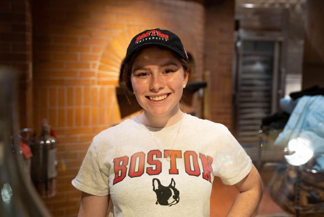 Photo: A young white woman stands in the middle of the photo, smiling, with a white t-shirt on, with red lettering that reads BOSTON. She has freckles. She is also wearing a black BU cap. She is in front of a brick background.