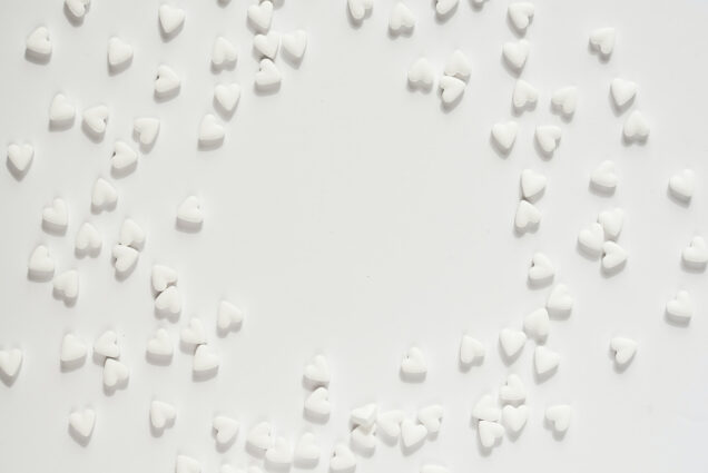 Photo: A bright overhead shot of a white table with scattered white heart-shaped Valentine's candies. Candies are semi-arranged so that there is a large space in the center.