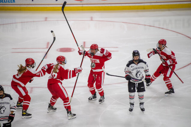 Photo: BU Women's team celebrates as they win a goal. They are wearing their red uniforms with white lining, their arms are open in celebration, there are four of them. Between the third and fourth BU player is a NE Huskies team member in a white and navy uniform. They are on an ice hockey rink.