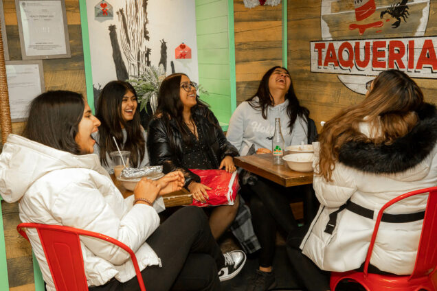 Photo: Five young feminine-presenting individuals laugh and dine at El Jefe's Taqueria. They sit at two, parallel tables. From left, the individual wears a white jacket and long black hair. Next, the individual has a light, gray shirt and long black hair. The next individual has square glasses, a black button up, and long black hair. They are throwing their head back to laugh. Second to last, on the right, the individual has a light gray hoodie on and long, black hair. The right-most individual has a white puffer coat on with black fur trim on the hood. Their hair is long, with black roots and bleached from the crown of their head, down.