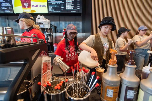 Photo: Students are pouring and making coffee at Saxby's coffee bar. They are wearing a red uniform. One individual is wearing a black bucket hat, a tan vest, and a cream shirt.