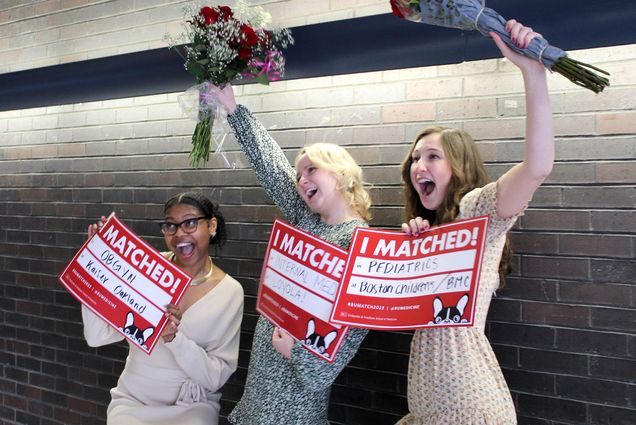 Photo: Three young women shout and raise their arms with glee. They're all dressed in business casual dresses and hold red signs and bouquets.