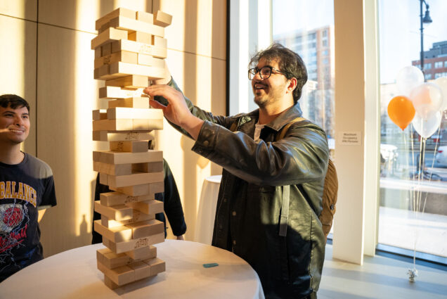 Photo: A tan young man wearing glasses and a leather jacket smiles as he carefully pulls out a wooden pice from a large Jenga tower. Another student smiles as they look on. Both stand in front of a large window letting it a sunset glow.