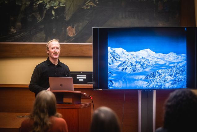 Photo: Griffin Post stands at a podium and looks to the audience. A white man wearing a black long-sleeved collared shirt stands in front of a podium where a silver laptop rests as he smiles and looks to the audience. To his right, a large screen shows a photo of a snowy mountain scene.