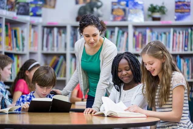 Photo: Elementary students are shown working with a teacher in library. A few small students look on as a teacher bends over them and points something out to them in a book on the table. Small bookcases can be seen behind them.