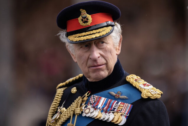 Photo: King Charles III looks to his right.An older white man wearing a traditional black military outfit and hat all brimming with gold chains, a row of various medals on the lapel, and various other gold and red embellishments, looks to his right.