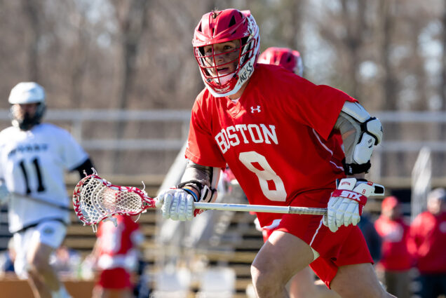 Photo: Captain Louis Perfetto, a BU men's lacrosse player in a red lacrosse uniform labeled "Boston 8" and helmet, runs as he holds a lacrosse stick on the field.