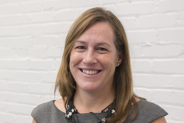 Photo: A white woman with shoulder length light, brown hair smiles in a portriat shot of her. She is wearing a big chunky necklace and a grey, mock-sleeve top.