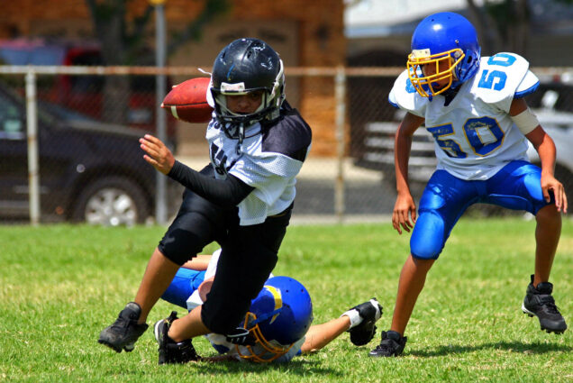 Photo: Young American football player in the running back position and wearing a black and white uniform is shown breaking away from an attempted tackle from another young player wearing a blue and white uniform. A third young player is shown in the same blue and white uniform to the right running alongside them.