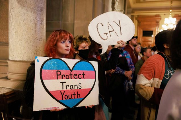 Photo: Protesters hold a placard that says, "Protect trans youth" and "Gay" inside the Indiana Statehouse. A young woman with short curly red hair holds a sign that reads "Protect trans youth" and a masked person next to her holds a word bubble sign that reads "Gay" . The two stand with other masked and unmasked protestors in a statehouse.