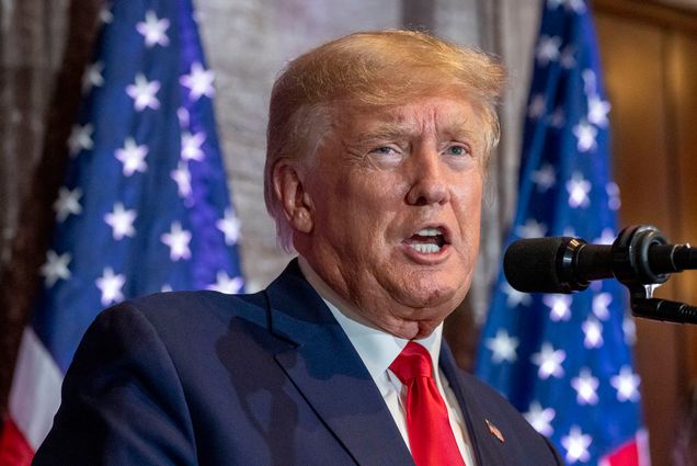 Photo: Candid photo of Donald Trump speaking. An older white man with an orange-tinged tan and wearing a navy suit and red tie speaks into a microphone. A US flag is seen behind him.