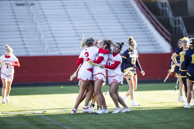 Photo: BU women lacrosse players celebrate after a goal against Merrimack in the 2023 season opener. A small group of young women wearing white and red lacrosse uniforms and sticks hug in the middle of a lacrosse field. A few young women dressed in navy and yellow lacrosse uniforms walk to their right.