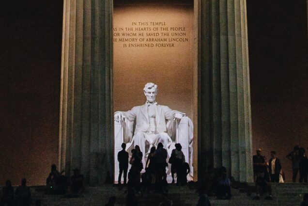 Photo: A night shot of the Lincoln Memorial, with the huge statue of President Lincoln looming the middle of the memorial. His statue is framed by two columns, vistors are in the foreground, shadowed by the night and a light shines on the inscription above the statue.