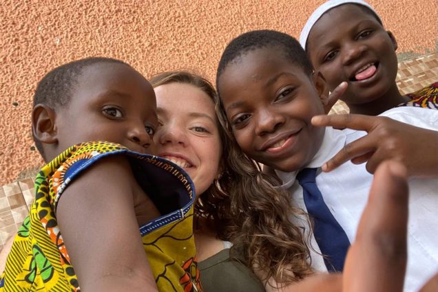 Photo: Tess Pollins, a white woman with short hair, with some of “her kids”, young black school children from Tanzania’s Chakuwama Orphanage, the beneficiary of her nonprofit. Photo courtesy of Tess Pollins