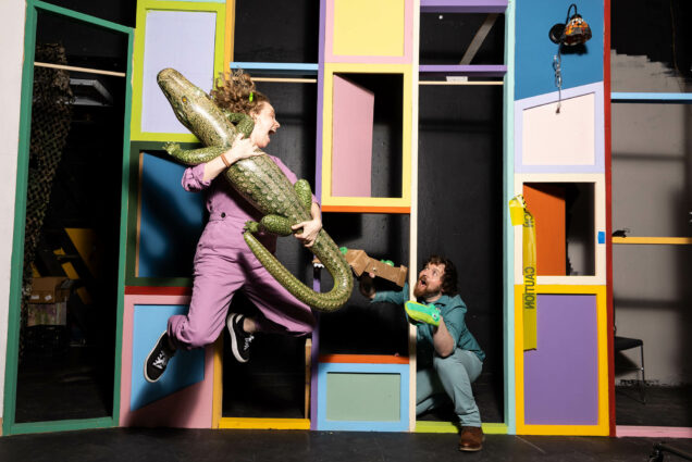 Photo: Jay Eddy (CGS’23) (left), the writer, composer and performer of Alligator-A-Phobia in 3D!, clowning around with director Shamus (CFA’23) on set. A woman with red hair and wearing a purple jump suit is shown mid-air as she jumps with a large, blow-up alligator in her arms. To he right, a man with dark brown curly hair and wearing a teal shirt and jeans peaks out from behind a set piece window. He has puppet alligators on his hands as looks to the woman to his left.