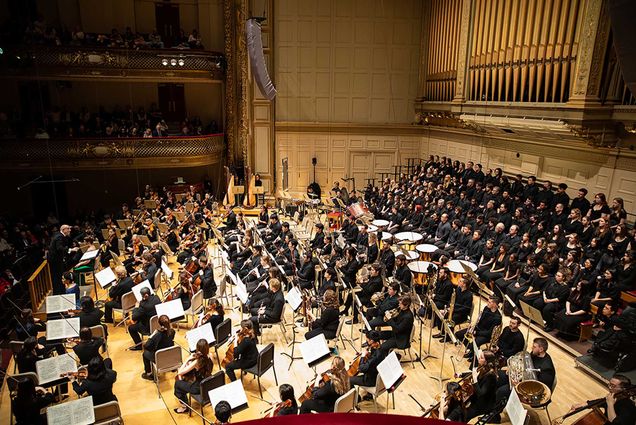 Photo: Overhead shot of a student symphony playing for a large audience in a symphony hall. Students wear all-black outfits as they play various instruments. A conductor stands to the left at the front of the orchestra, as his back faces the audience behind him.