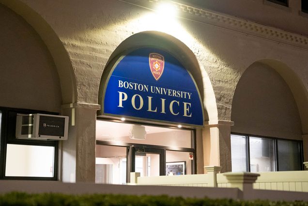 Photo: Exteriors photo of the Boston University Police Department. A white brick building is shown at night illuminated by a bright spotlight over the front door. A blue sign over the door reads "Boston University Police".