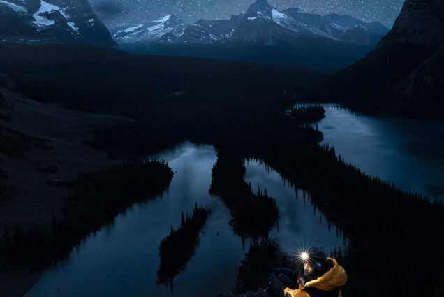 Photo: Large, dramatic photo of a starry night sky over scenic mountains. A person can be seen in the foreground wearing a yellow puffer jacket and a headlamp that illuminates him and the area around him. Bodies of water can be seen around dark trees below the cliff the man sits on.