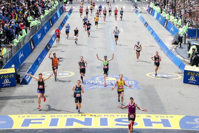 Photo: Runners cross the finish line at The Boston Marathon in 2022. There are large crowds gathered on the sides of the race course and more than 10 runners are nearing the large yellow finish line.