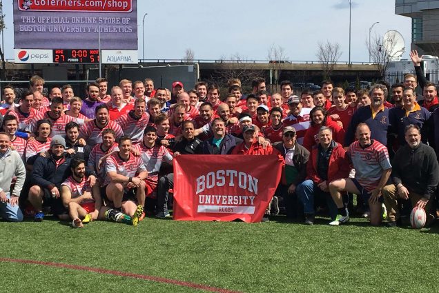Photo: A group photo of BU’s Men’s Club Rugby Team during their 45th anniversary. A group of men, many wearing red Rugby jackets, pose together on Nickerson soccer field. A few people in the front hold up a large red banner that reads "Boston University Rugby".