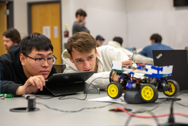 Photo: Yang Yang Zhang (ENG’24) (left), and Kai Imery (ENG’24) work to get their object-detecting autonomous vehicle to run. A young Asian man wearing glasses and a young white man wearing a white hooded sweatshirt both lean close the table in front of them as they study the small electric car they're programming. The small blue car is connected to wires and attached to a small black device that the two are working on.