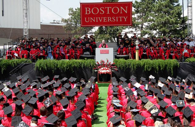 This is a birds eye view photo from a Boston University commencement ceremony. Seniors are wearing black graduation caps with red gowns and are sitting down in rows. The stage is in front of the graduates and there is someone shown speaking at the podium. Behind the speaker, there are rows of faculty/speakers/staff also in red and black graduation attire.