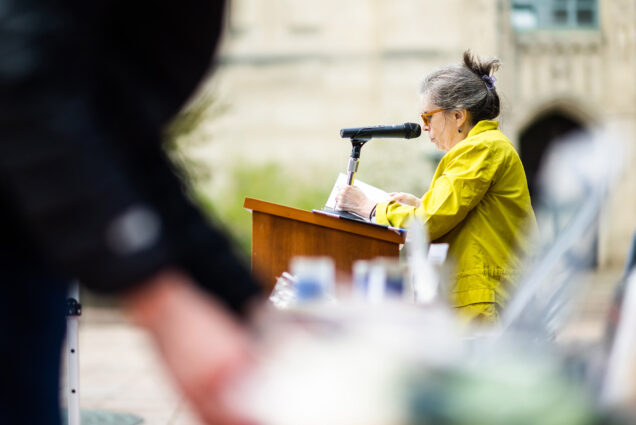 Photo: An older woman with graying black hair tied up and wearing a bright yellow jacket stands at a podium outside in an open square and reads from a large packet of papers. In the blurry foreground, a hand can be seen picking up a newspaper.