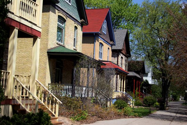 Photo: A sidewalk lined with cookie-cutter houses on the left side. They are tan with red, or blue roofs. Most have a porch with an awning on it. There are trees lining the other side of the sidewalk. The day is bright and sunny.