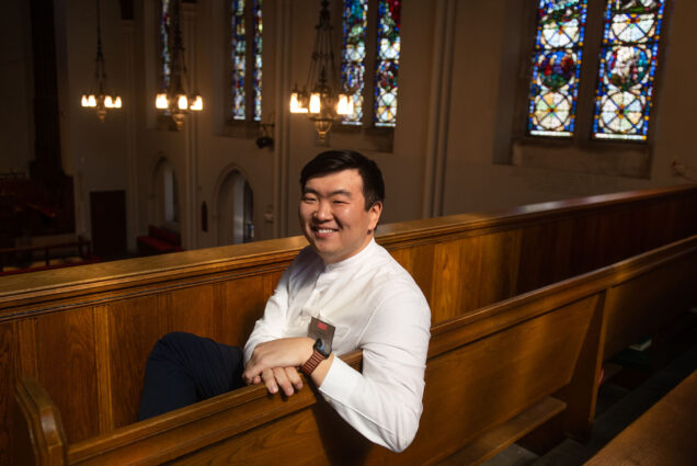 Photo: Jonathan Lee, an Asian man wearing a white long-sleeved chaplain shirt, black pants, and a silver name tag that reads "Jonathan Lee" sits posed in a pew. He smiles, as he looks over his left shoulder to the camera in a church.