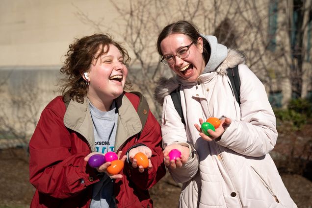 Photo: Two young women in winter gear laugh as they lean towards each other and display colorful, plastic Easter eggs in their hands towards the camera.