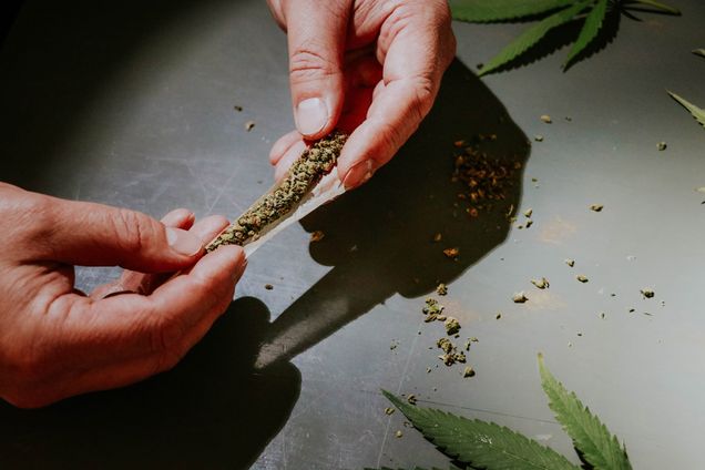 Photo: Overhead shot of a person's hand as the roll a joint of weed. A dried plant is being rolled between brown paper of a table in a darkened room.