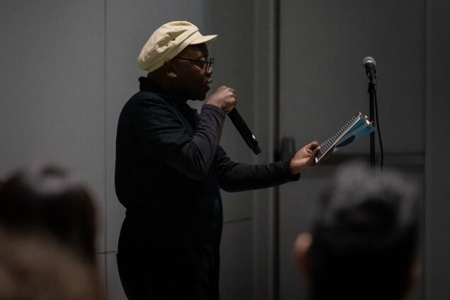 Photo: Award winning Brazilian Poet and performer, Ricardo Aleixo, a Black man wearing an all black outfit and tan beret-style cap holds a microphone to his face as he reads from a small notebook. A mic stand is seen to his right and blurred views of the back of audiences heads can be seen in the foreground.
