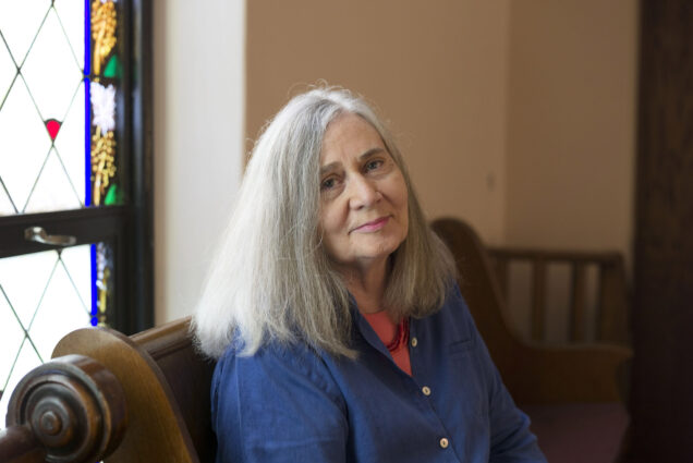 Photo: A portrait of Marilynne, a white woman with shoulder length gray hair. She is wearing a deep blue blazer and sits on a brown leather couch with a stained-glass-trimmed window behind her. She smiles softly for the camera.