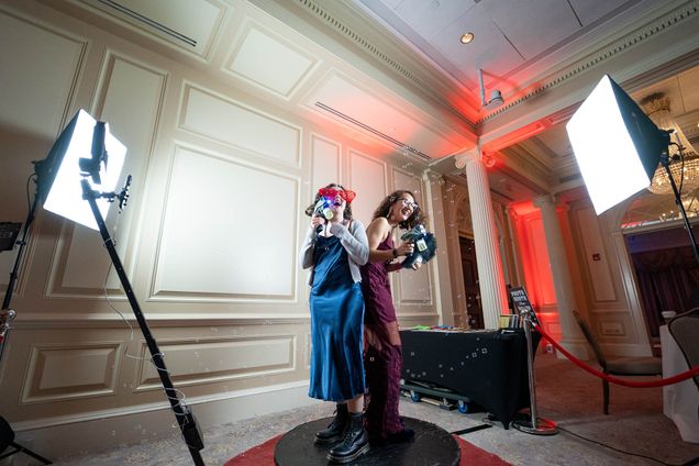 Photo: Danielle Miller (CAS'24) and Elizabeth Nelson (CAS'23) (Left to right) use the photo booth at the Spring 2023 Questrom Formal QSG hosted by Questrom Student Government. Two young women, (from left) one wearing a shiny, satin blue dress with Doc Martin boots and the other wearing long, burgundy dress laugh and pose with funky props like large, red clown glasses in front of white wall. The photo booth station features large light screens and pedestal stand.