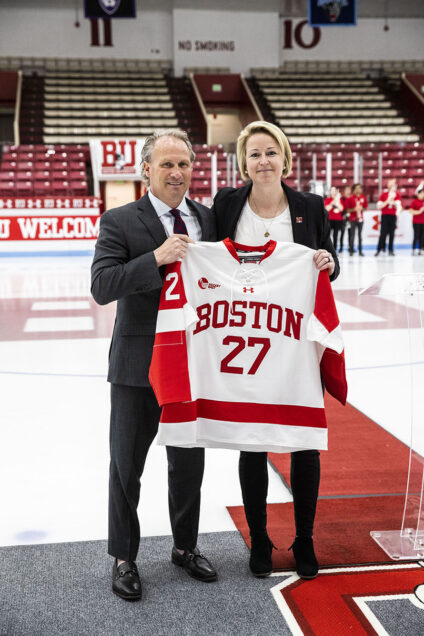Photo: Tara Watchorn, a white woman with short blonde hair, and Drew Marrochello, a white man with silver hair, stand on a red rug on an ice rink as they pose together and hold up a large BU women's ice hockey jersey. They both wear formal black blazers, white tops, and black pants. Behind them empty stands can be seen and students in the BU band wait with their instruments in the distance.
