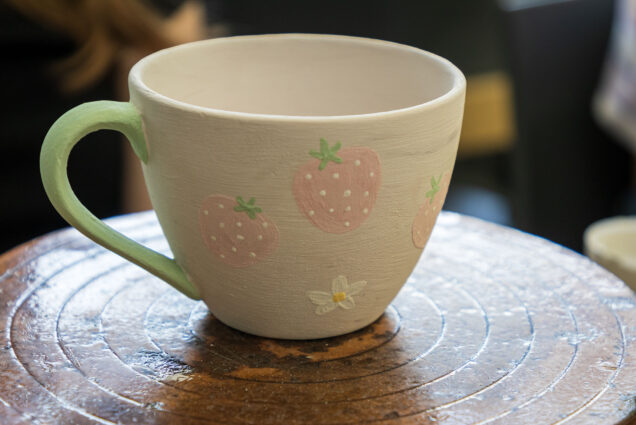 Photo: A picture of a ceramic mug before it goes in the kiln. It has green glaze on the handle and little strawberries and little white flowers.