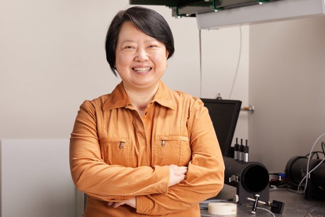 Photo: Xin Zhang, an Asian woman with short black hair and wearing a burnt orange collared shirt is shown smiling and standing with arms crossed poses in her lab. She wears Computer materials can be seen behind her.