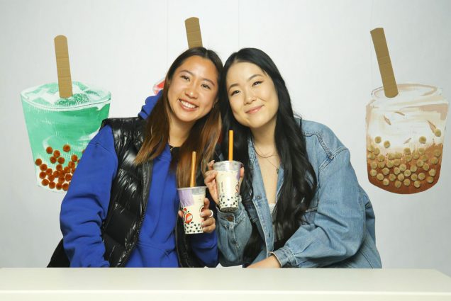 Photo: Sharlene Chang (SHA’24) and Irene Kim (COM’23), two young Asian women holding boba teas, smile and pose resting together. They sit in front of a backdrop of large boba tea recreations made out of cardboard and puff balls.