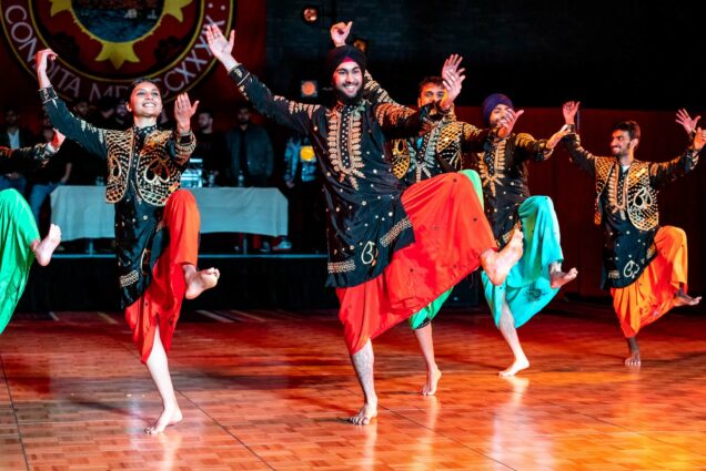 Photo: The BU Bhangra dance team celebrates Punjabi Nite at BU with a colorful and lively performance. The dancers are smiling, their arms in the air and a foot of the ground, mid-dance. They wear traditional clothing--a black embellished tunic with various colorful parachute-type pants on the bottom. The colors range from red to a blue-green, left to right.