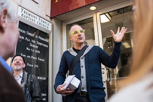 Photo: Journalist Scott Kirsner, a white man wearing neon yellow glasses, a black shirt, navy blue and grey button-up sweater, and jeans holds his hand up as he talks to a crowd. He clutches a few miscellaneous items in his other hand as he stands in front of glass doors to a building. The sign to his left reads "Tremont Temple Baptist church". Another man to his left smiles and laughs.