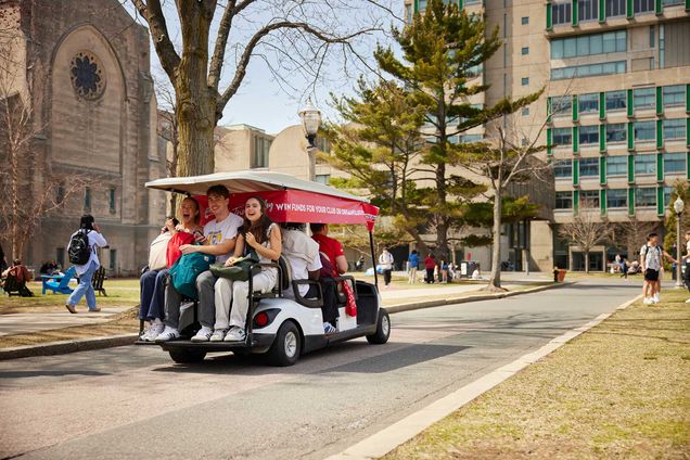 Photo: Three kids sit paced onto the back of a trolley zooming through BU campus. The day is sunny, the individuals are smiling, and the trolley cart is in the BU colors--red and white.