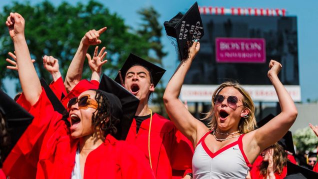This is photo taken at a past Boston University commencement ceremony. It is outdoors on a sunny day, with students in their caps and gowns shown, cheering with their arms up. Photo Credits: Janice Checchio