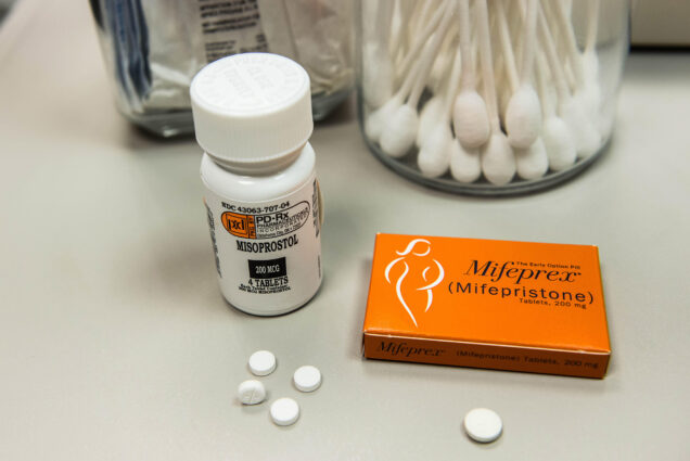 Image: A stock photo of Mifepristone. To the right is a bottle of Mifepristone and to the left, a orange box containing Mifepristone. In front of both are four scattered pills of Mifepristone. Behind them, miscellaneous items found in a doctors office such as a jar of Q-tips and a jar of something that is single-packaged.