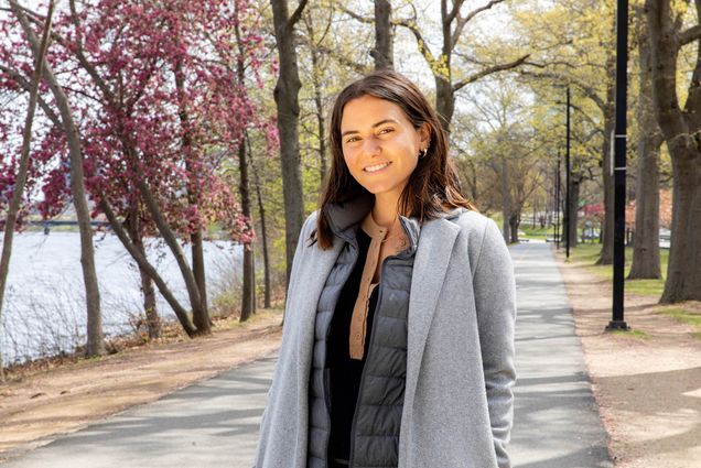 Photo: A hispanic young woman, Paulina Preciat Jasso, stands outside on a sunny day in a gray jacket and black and tan top. She has shoulder length brown hair and smiles for the camera.