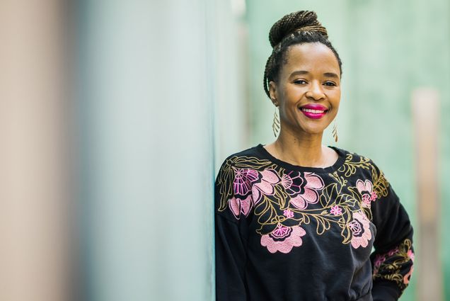 Photo: A portrait of Thato Mwosa, a Black women with braids tied up in a bun on the top of her head. She smiles for the camera while wearing a black shirt with floral embroidery.